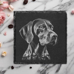 May include: A square, black slate coaster with a detailed, grayscale portrait of a dog. The dog has a focused gaze and a sleek coat. The coaster is surrounded by pink and yellow flowers, coffee beans, and a marble surface.