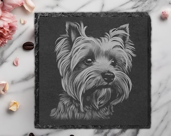 May include: A black slate coaster with a detailed engraved image of a Yorkshire Terrier dog. The dog is looking directly at the viewer with a serious expression. The coaster is surrounded by a white marble surface with pink flowers and coffee beans.
