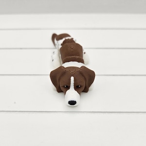 May include: A brown and white 3D printed dog figurine with a white face and brown ears. The dog is lying down with its head resting on the ground.