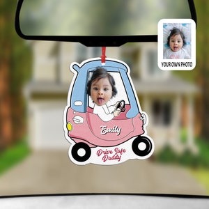 Puede incluir: Un ambientador de coche personalizado colgado del retrovisor, con un coche de juguete rosa y azul y la foto de un beb&eacute; dentro. El nombre Emily aparece en el lateral y el texto Drive Safe Daddy est&aacute; escrito en rojo en la parte inferior.