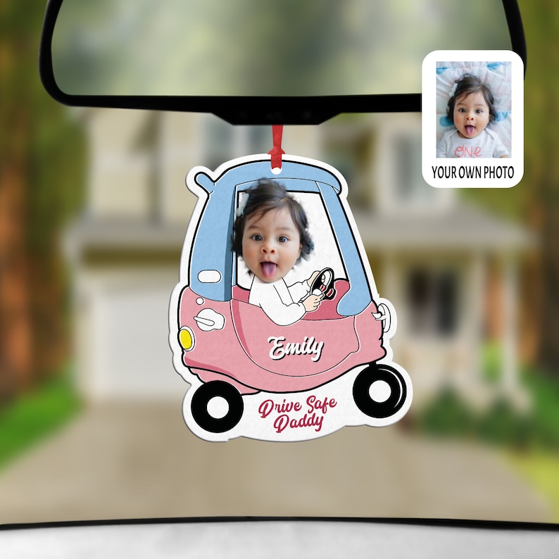 Puede incluir: Un ambientador de coche personalizado colgado del retrovisor, con un coche de juguete rosa y azul y la foto de un beb&eacute; dentro. El nombre Emily aparece en el lateral y el texto Drive Safe Daddy est&aacute; escrito en rojo en la parte inferior.