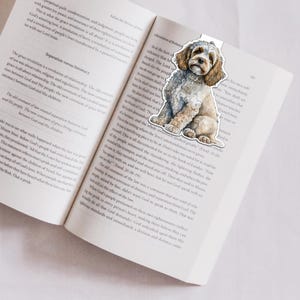 May include: A bookmark with a watercolour illustration of a fluffy, light brown and white dog. The bookmark is on an open book with printed text. The dog is sitting and looking forward.