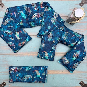 May include: A set of blue fabric heating pads with a feather pattern in shades of teal, gold, and red. The pads are arranged on a light blue wooden surface. A small glass jar with a white lid is also visible.