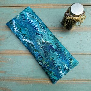 May include: A rectangular, blue and teal patterned fabric item with a marbled design. The fabric is folded and lies on a weathered, turquoise wooden surface. A small glass jar with a silver lid and dried grass tied around it is in the background.