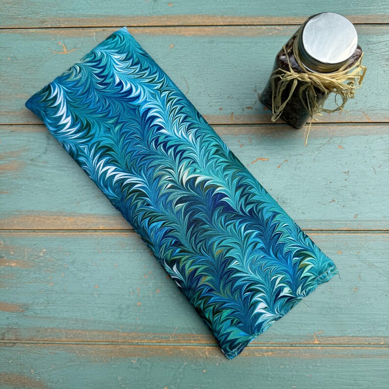 May include: A rectangular, blue and teal patterned fabric item with a marbled design. The fabric is folded and lies on a weathered, turquoise wooden surface. A small glass jar with a silver lid and dried grass tied around it is in the background.