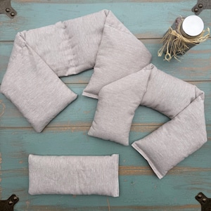 May include: A collection of gray fabric heating pads in various shapes, including a long, segmented wrap and a rectangular pad. The pads are made of a soft, textured material and are arranged on a weathered, teal-colored wooden surface.