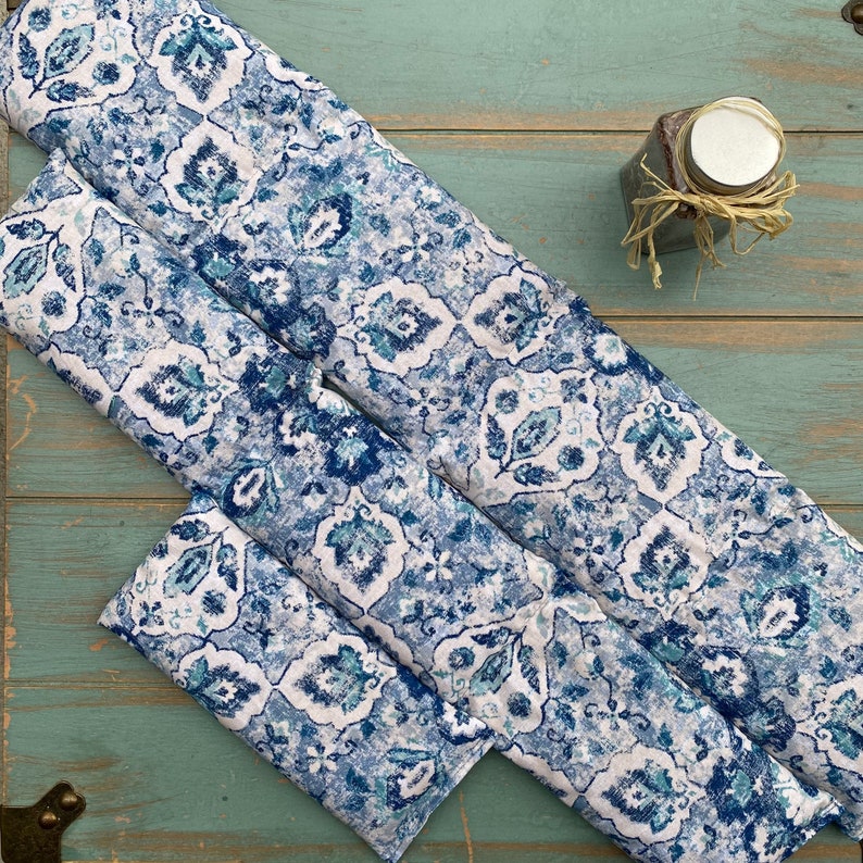 May include: Three blue and white patterned fabric eye pillows. The pillows are rectangular and have a floral design. The pillows are arranged on a light blue wooden surface.