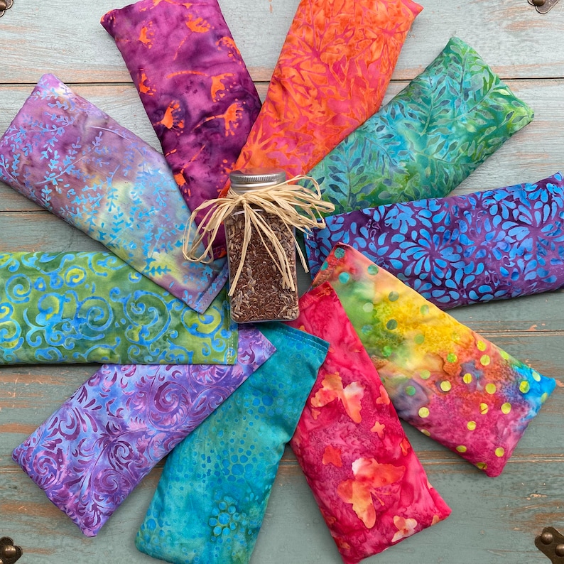 May include: A collection of colorful fabric eye pillows with a glass jar of dried herbs in the center. The eye pillows are made of different patterns and colors, including blue, green, purple, red, and orange.
