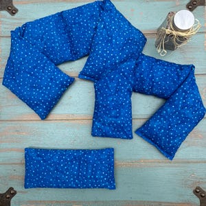 May include: A collection of blue fabric heat packs with white polka dots. The packs are in various shapes and sizes, including long, rectangular, and square. The fabric has a textured appearance. A small jar with a lid is also visible.