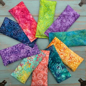May include: A collection of ten colorful eye pillows arranged in a circle. The pillows are made of fabric with various patterns, including floral, geometric, and abstract designs. The pillows are all rectangular in shape and are approximately 6 inches long and 3 inches wide.