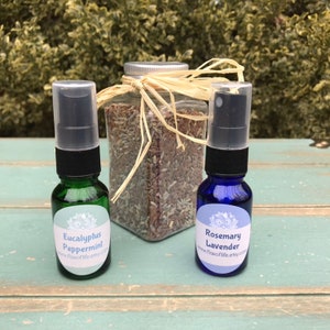 May include: Two small glass spray bottles with labels. The bottle on the left is green and says "Eucalyptus Peppermint" and the bottle on the right is blue and says "Rosemary Lavender". Both bottles have a clear spray nozzle and a black cap. A glass jar filled with dried lavender is behind the bottles.