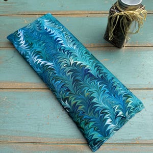May include: A rectangular, blue and teal fabric item with a marbled pattern. The fabric is filled and appears to be a heat pack or similar item. A small glass jar with a metal lid and twine is in the background.