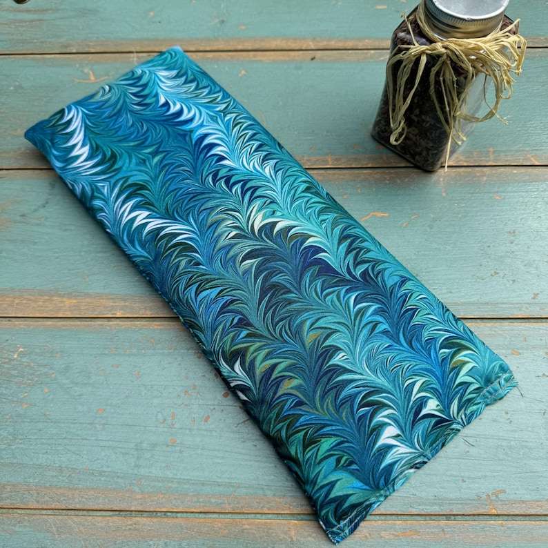 May include: A rectangular, blue and teal fabric item with a marbled pattern. The fabric is filled and appears to be a heat pack or similar item. A small glass jar with a metal lid and twine is in the background.
