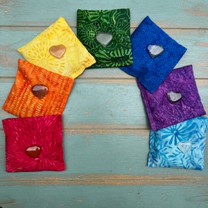 May include: Seven colorful fabric pouches arranged in a rainbow pattern, each containing a heart-shaped stone. The pouches are made of different patterned fabrics and are sewn together to form an arch.