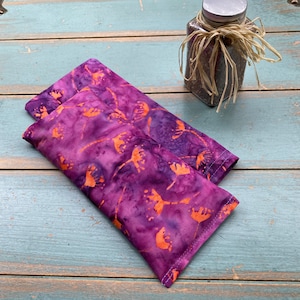 May include: Two purple and orange patterned eye pillows. The pillows are made of fabric with a batik design. The pillows are lying on a blue wooden surface.