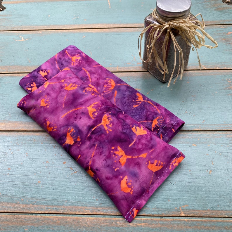 May include: Two purple and orange patterned eye pillows. The pillows are made of fabric with a batik design. The pillows are lying on a blue wooden surface.