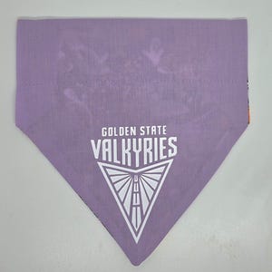 May include: A triangular lavender fabric bandana featuring the white text GOLDEN STATE VALKYRIES above a white graphic of a stylized downward-pointing triangle containing a road-like design with vertical lines.