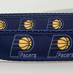 May include: A navy blue dog collar featuring the Indiana Pacers basketball team logo. The collar has a repeating pattern of yellow basketballs and the team's logo. It includes a silver metal D-ring and a blue plastic buckle.