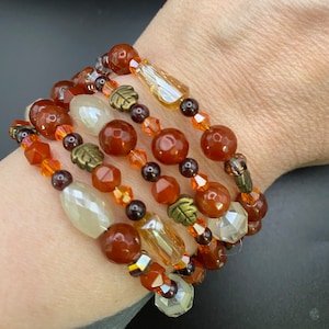 May include: A set of five bracelets featuring carnelian, clear and amber glass beads, and gold-toned leaf charms. The bracelets are layered on a person's wrist.