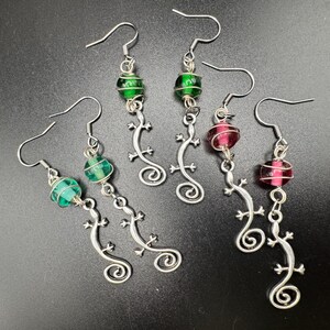 May include: Earrings with silver-tone lizard charms and colorful glass beads. The earrings feature green and red beads, with a coiled tail design. The hooks are silver-colored. The earrings are designed for pierced ears.