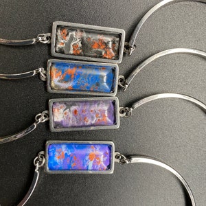 May include: Four silver bracelets with rectangular resin charms. Each charm features a different abstract design with blue, purple, and orange colors.