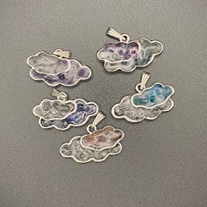 May include: Five cloud-shaped pendants with silver-colored trim. Each pendant features a unique color scheme, including shades of purple, blue, and brown. The pendants have a bail for attaching to a chain or cord.