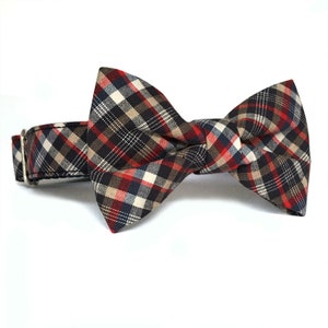 May include: A red, black, and tan plaid bow tie for a pet collar. The bow tie is made of fabric and has a classic bow shape.