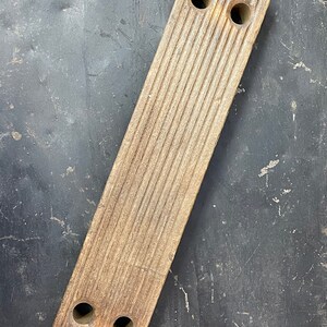 May include: A long, rectangular wooden block with a grooved surface and four holes drilled through it. The block is a light brown color and has a worn, rustic appearance.