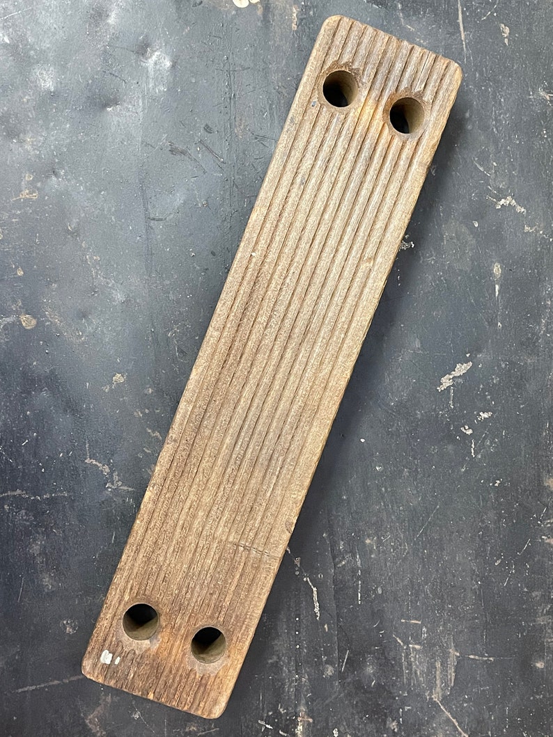 May include: A long, rectangular wooden block with a grooved surface and four holes drilled through it. The block is a light brown color and has a worn, rustic appearance.