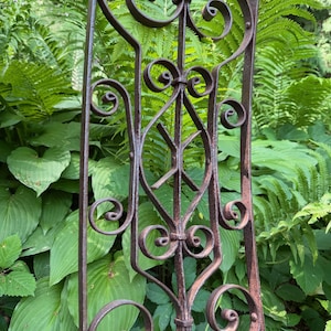 May include: A tall, rectangular wrought iron panel featuring a symmetrical design of scrolled metalwork and a central diamond-shaped frame. The metal has a weathered, dark brown, rusted finish with visible signs of oxidation throughout the structure.