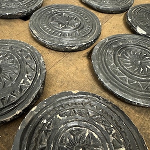 May include: A collection of dark gray, round, carved stone coasters with intricate floral and geometric designs. The coasters have a distressed, aged appearance with white accents, resting on a wooden surface. The coasters are approximately 4 inches in diameter.