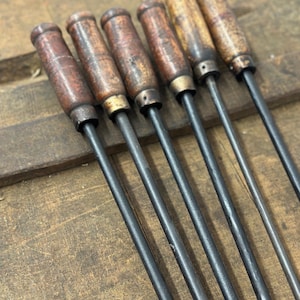 May include: Six vintage metal skewers with wooden handles. The handles are a dark brown color with a worn, aged appearance. The metal shafts are dark gray and appear to be made of iron. The skewers are arranged diagonally across a wooden surface.