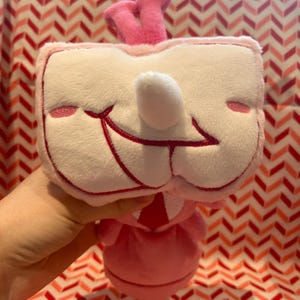May include: A plush toy in the shape of a smiling face with a white nose and pink accents. The toy has a pink crown and a pink dress. The background is a red and white chevron pattern.