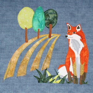 以下が含まれることがあります： 風景の中にキツネを描いたテキスタイルアート。キツネはオレンジ、白、グレーで表現されています。緑と黄色の葉を持つ3本の木と、曲線のあるフィールドが含まれています。背景は青色の生地です。