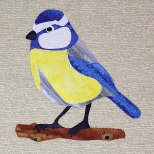 Puede incluir: Un pájaro azul con el pecho amarillo y las alas grises posado en una rama marrón. El pájaro está hecho de tela y tiene un diseño cosido.