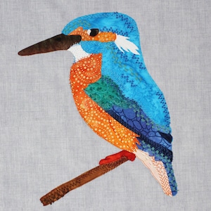 May include: A blue and orange patchwork bird with a long beak perched on a brown branch. The bird is made of fabric and has a detailed design.