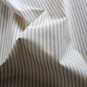 May include: Close-up of folded fabric featuring a cream background with vertical tan stripes. The material has a soft, textured appearance, with evenly spaced stripes. The folds create shadows and depth, highlighting the fabric's texture.