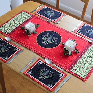 May include: A Christmas-themed table setting with a red runner and matching placemats. The runner and mats feature embroidered reindeer and Christmas tree designs. Silverware and candles add to the festive atmosphere.