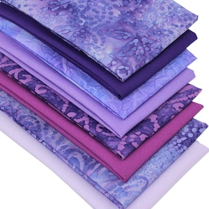 May include: A collection of fabric squares in shades of purple, from pale lavender to dark violet. Some fabrics have patterned designs, others are plain. The fabrics are stacked, displaying a gradient of colours.