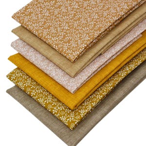 May include: A stack of folded fabric pieces in various shades of mustard yellow, tan, and light pink. Some pieces feature a white vine and floral pattern, while others are solid-coloured with a textured, woven appearance stacked in layers.