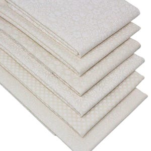 May include: A stack of six folded fabric squares in a light cream colour. Each piece of fabric features a distinct white tonal pattern, including floral motifs, small geometric shapes, and delicate leaf designs, all arranged in a neat diagonal pile.