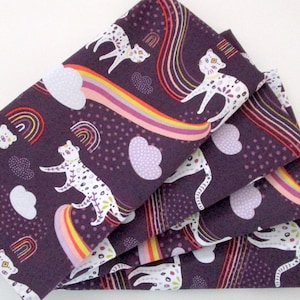 May include: Folded fabric with a whimsical pattern featuring white cats with floral markings, rainbows, clouds, and hearts on a dark purple background. The fabric is folded in a stack, showcasing the repeating design.