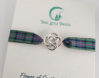 May include: A silver Celtic knot charm on a green, blue, and red tartan ribbon. The ribbon is attached to a white card with the text "Three Little Pirates" and "Flower of Scotland Tartan".