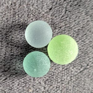 May include: Three round, sea glass pieces in shades of blue-green and green. The pieces have a frosted appearance and are arranged on a textured, gray surface. The sea glass is smooth and appears to be well-worn.