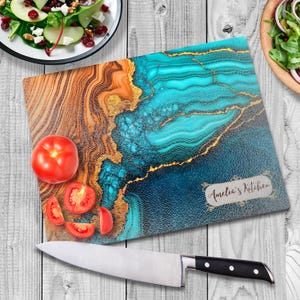 May include: A blue and gold abstract design glass cutting board with the text "Amelia's Kitchen" in a white rectangle. The board is on a white wooden surface with a knife and tomatoes.