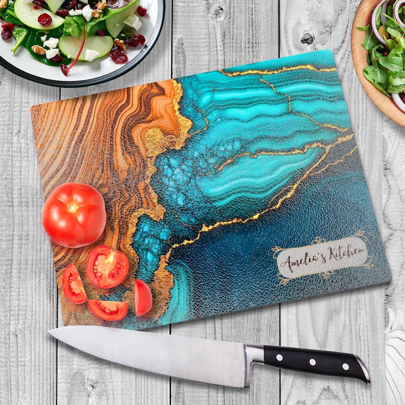 May include: A blue and gold abstract design glass cutting board with the text "Amelia's Kitchen" in a white rectangle. The board is on a white wooden surface with a knife and tomatoes.