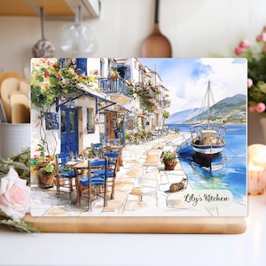 Könnte beinhalten: Ein rechteckiges Glas-Schneidebrett mit einem Aquarellgemälde eines Küstendorfes. Das Bild zeigt blaue Türen, Tische und ein Segelboot. Der Text "Lily's Kitchen" befindet sich unten rechts. Die Szene spielt vor blauem Meer und Himmel.