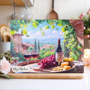 May include: A decorative cutting board with a painting of a Tuscan scene, including wine, grapes, and cheese. Two wine glasses are filled with red wine, and a bottle of wine is also present. The cutting board has the text "Lily's Kitchen".