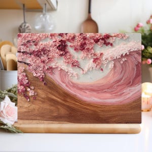 Pode incluir: Uma tábua de corte retangular de madeira com um desenho de flor de cerejeira. O desenho apresenta flores cor-de-rosa e um padrão de ondas em tons de rosa e branco. A tábua tem uma base de madeira.