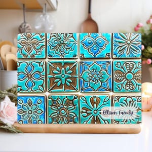 May include: A set of 12 turquoise and gold ceramic tiles with intricate floral patterns. The tiles are arranged in a grid pattern and have a glossy finish. The tiles are labeled "Ellison Family".
